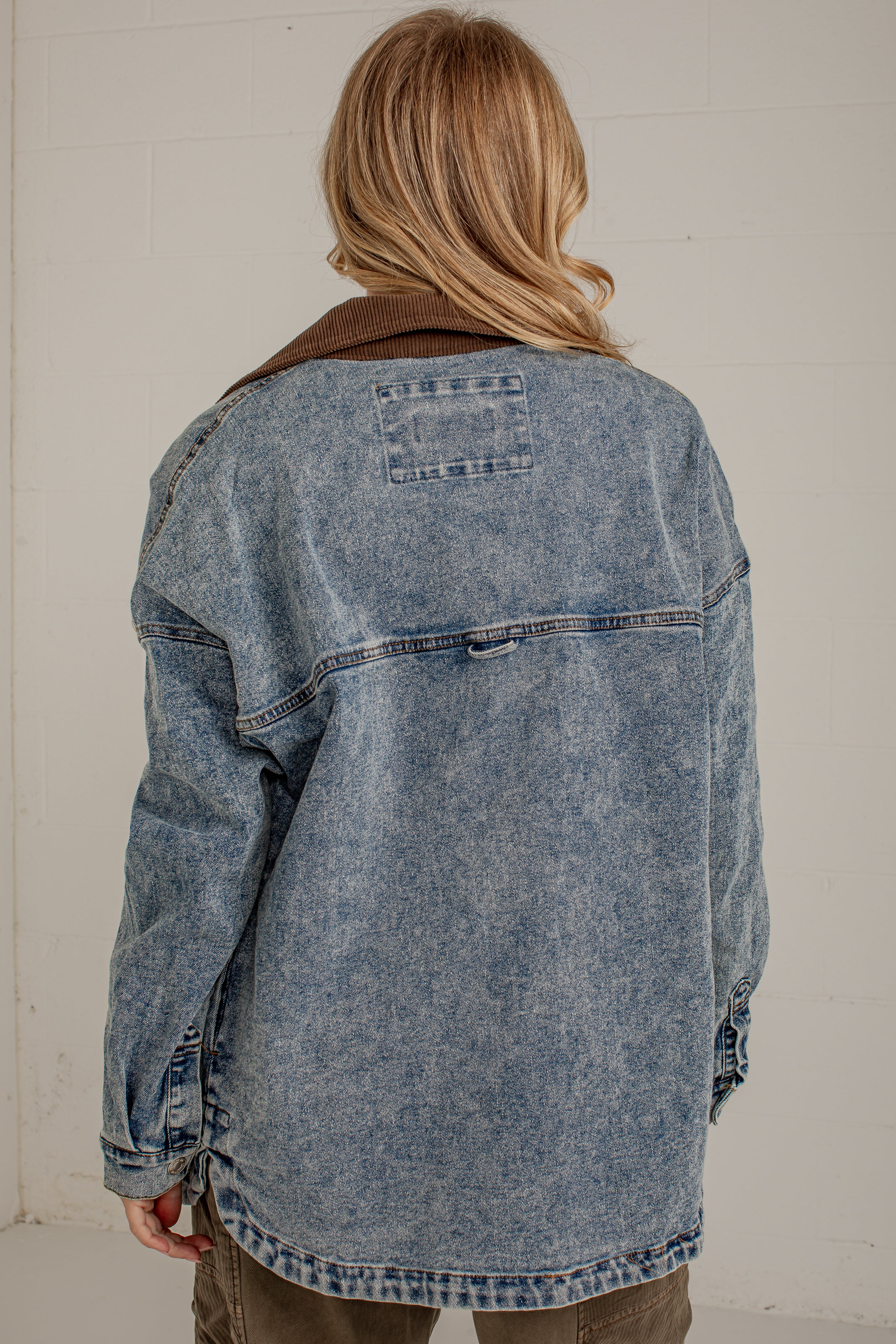 A person viewed from behind wearing an oversized washed blue denim jacket with a brown corduroy collar, paired with olive pants, against a light off-white brick wall.