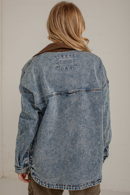 A person viewed from behind wearing an oversized washed blue denim jacket with a brown corduroy collar, paired with olive pants, against a light off-white brick wall.
