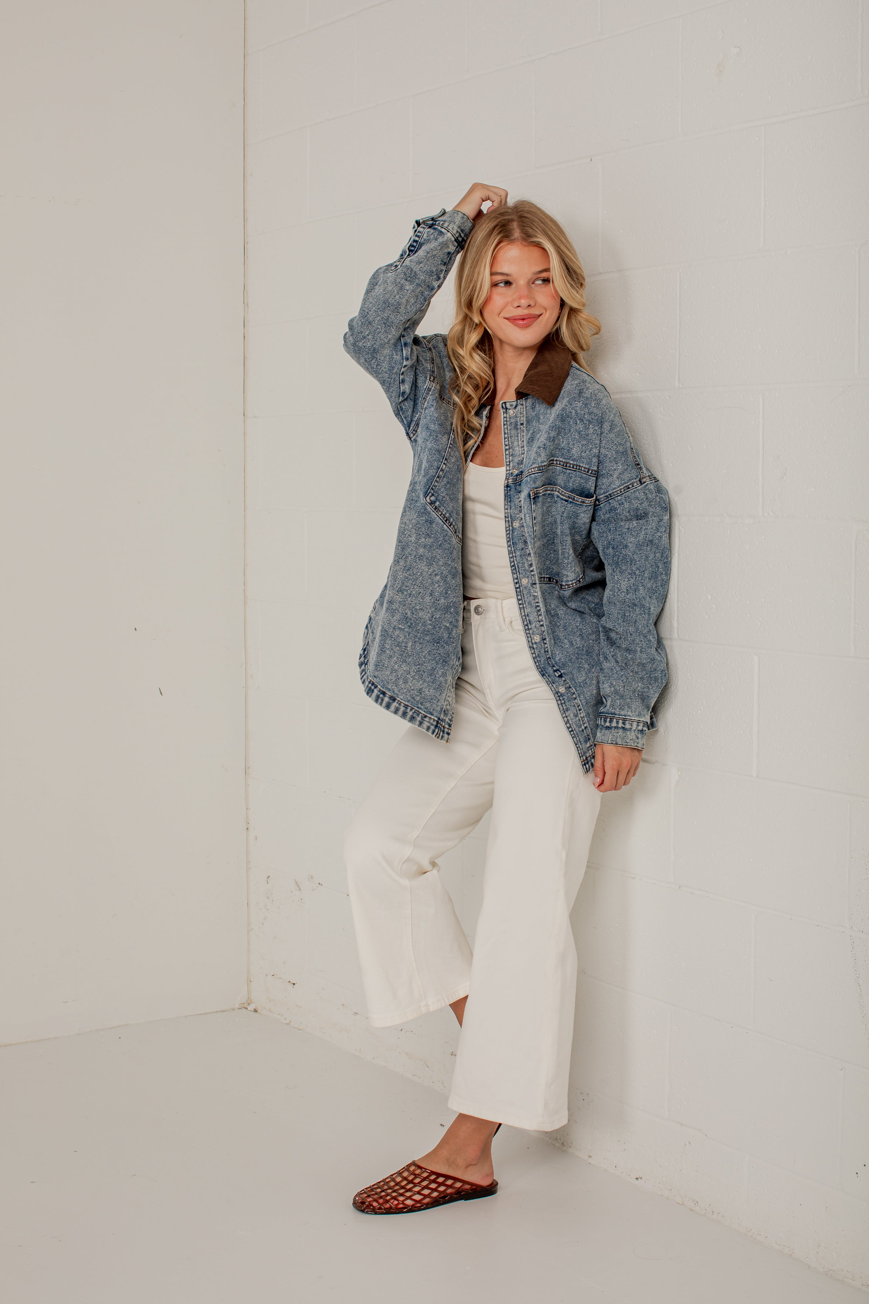 A woman models a washed blue denim jacket with a brown collar, paired with white wide-leg pants and brown woven mule flats, posing in a minimalist white room.