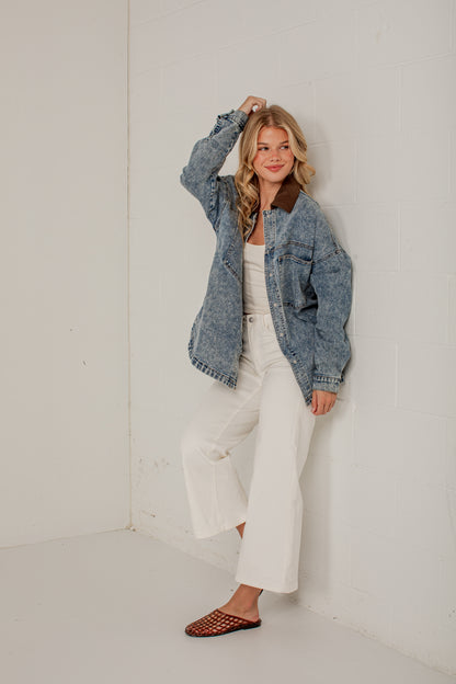 A woman models a washed blue denim jacket with a brown collar, paired with white wide-leg pants and brown woven mule flats, posing in a minimalist white room.