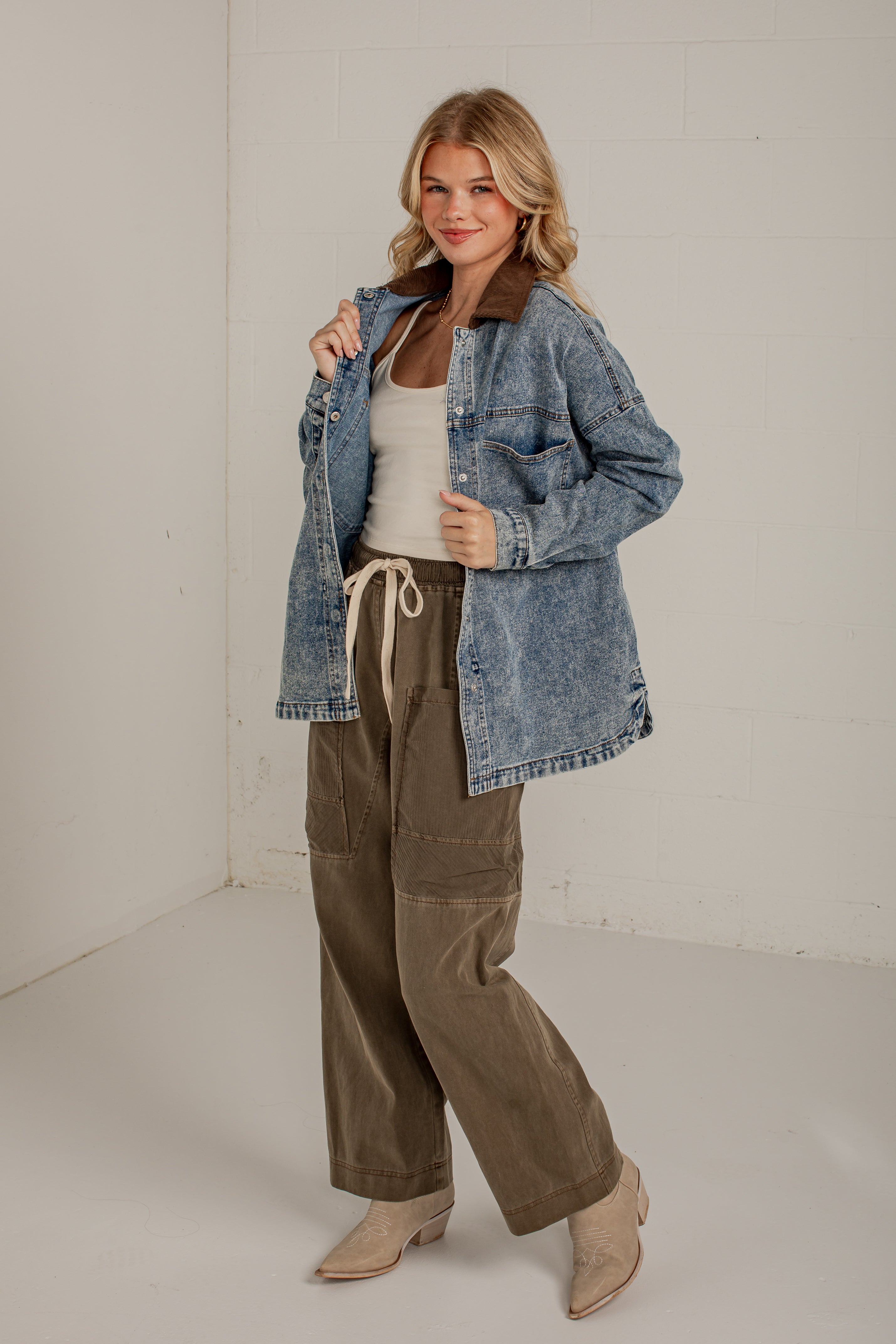 A woman modeling a light blue washed denim jacket with a brown corduroy collar, worn over a white tank top and olive green wide-leg pants, in a bright studio setting.