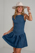 A woman wearing a sleeveless smocked blue dress and a wide-brimmed beige hat, posing in front of a white brick wall.