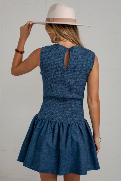 A woman wearing a sleeveless blue smocked dress with a flared skirt, shown from the back while wearing a wide-brimmed cream hat against a white brick wall.