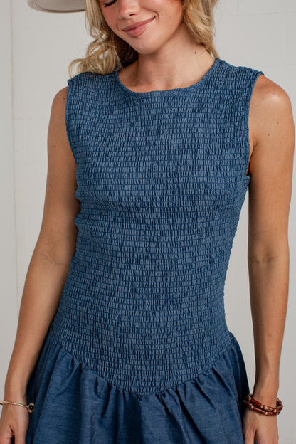 A woman wearing a sleeveless smocked blue dress, cropped from the chin down, against a plain light background.