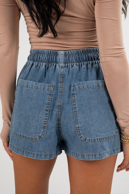 Person wearing blue denim shorts and a beige long-sleeve top on a plain background