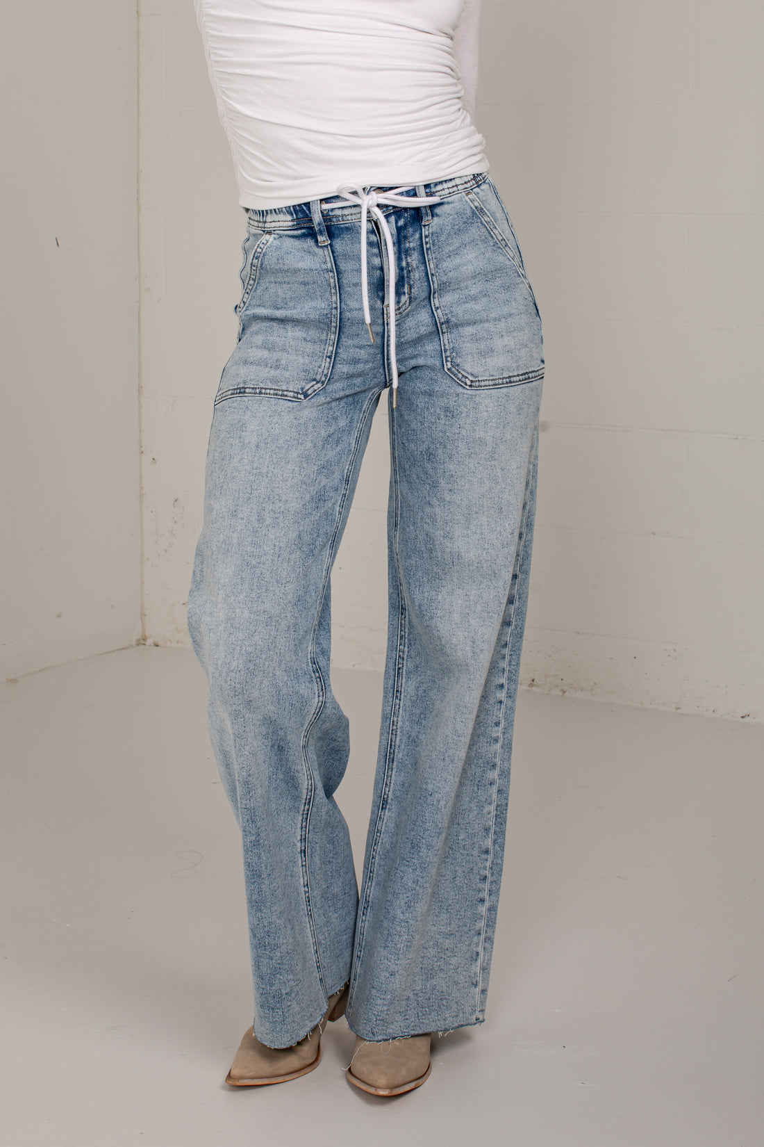 A person wearing light-wash wide-leg denim jeans with a drawstring waist, paired with a white top and tan pointed shoes, standing in a plain indoor studio corner.