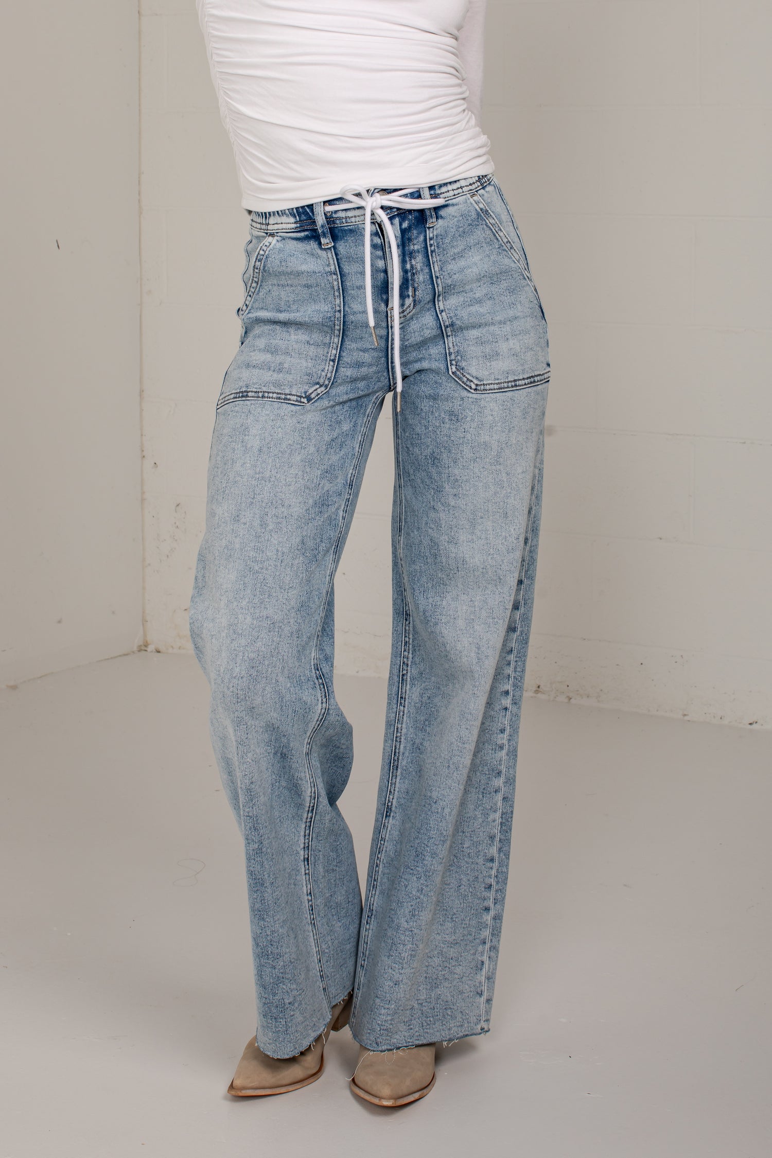 A person wearing light-wash wide-leg denim jeans with a drawstring waist, paired with a white top and tan pointed shoes, standing in a plain indoor studio corner.