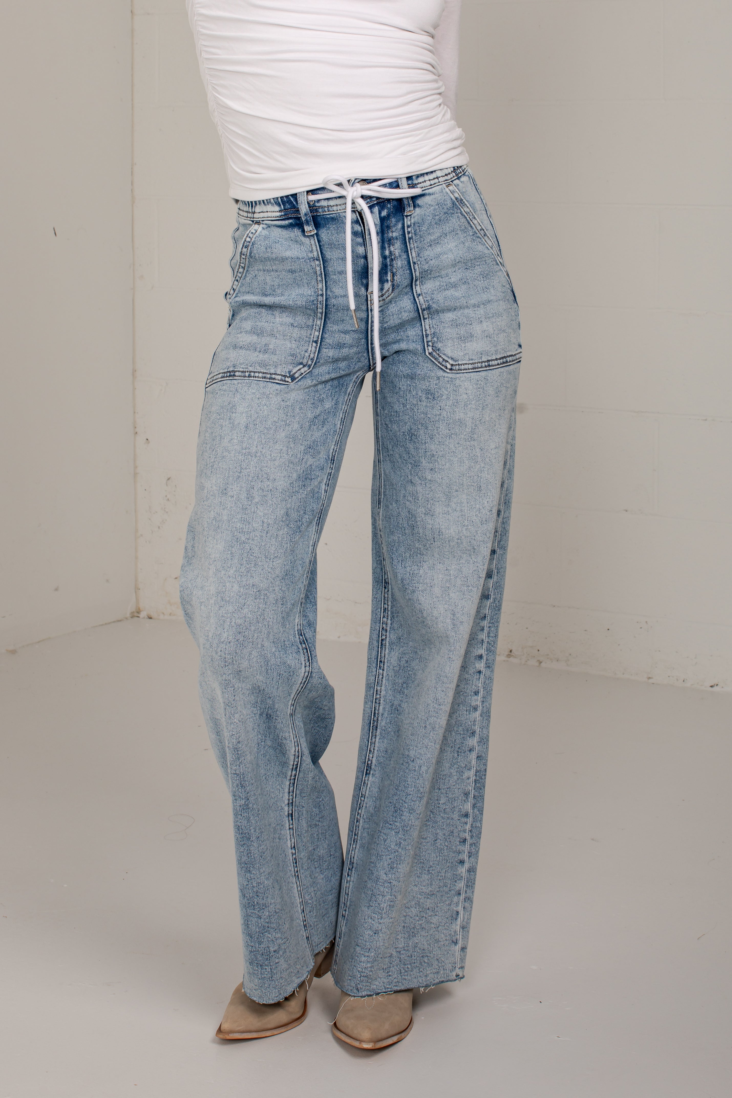 A person wearing light-wash wide-leg denim jeans with a drawstring waist, paired with a white top and tan pointed shoes, standing in a plain indoor studio corner.