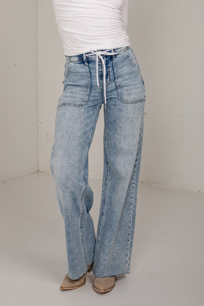A person wearing light-wash wide-leg denim jeans with a drawstring waist, paired with a white top and tan pointed shoes, standing in a plain indoor studio corner.
