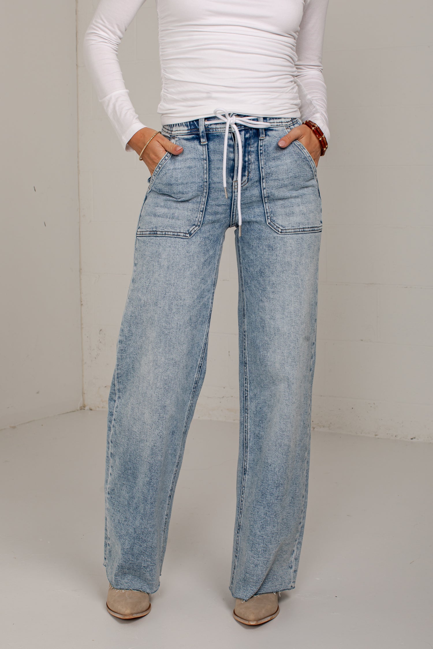 A person wearing light-wash blue wide-leg denim jeans with a white drawstring waist, paired with a white long-sleeve top and beige shoes, standing against a plain off-white wall.