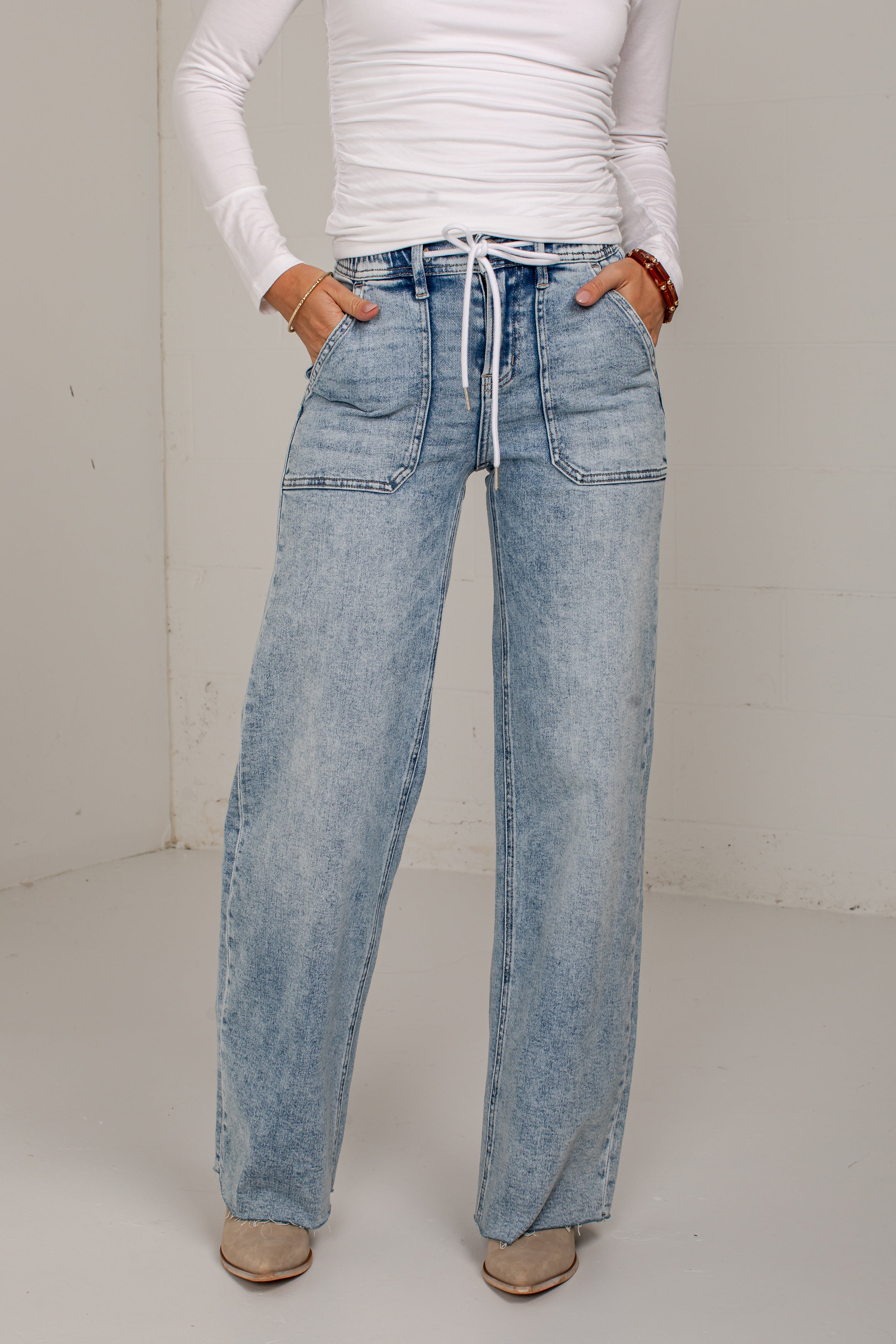 A person wearing light-wash blue wide-leg denim jeans with a white drawstring waist, paired with a white long-sleeve top and beige shoes, standing against a plain off-white wall.