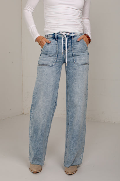 A person wearing light-wash blue wide-leg denim jeans with a white drawstring waist, paired with a white long-sleeve top and beige shoes, standing against a plain off-white wall.