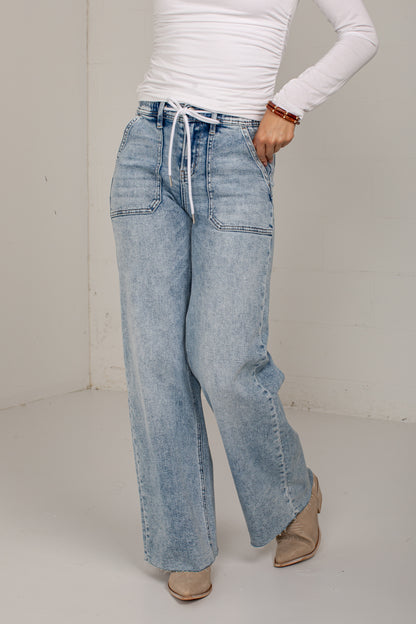 A person wearing light-wash wide-leg denim jeans with a white drawstring waist, paired with a white long-sleeve top and tan pointed-toe flats, standing in a plain indoor studio setting.