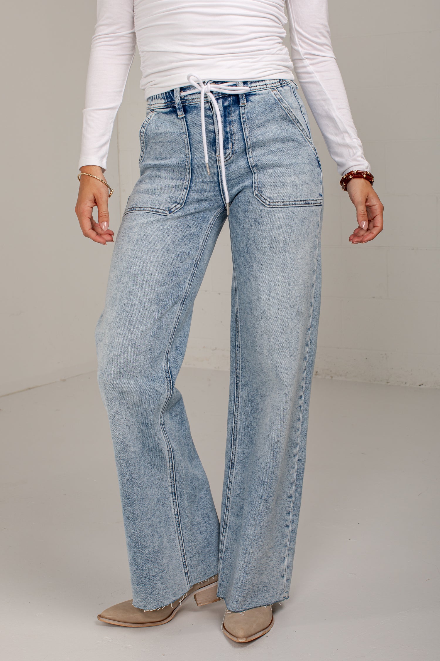 A person wearing light-wash wide-leg denim jeans with a drawstring waist, paired with a white long-sleeve top and beige pointed-toe heels, photographed against a plain light studio background.