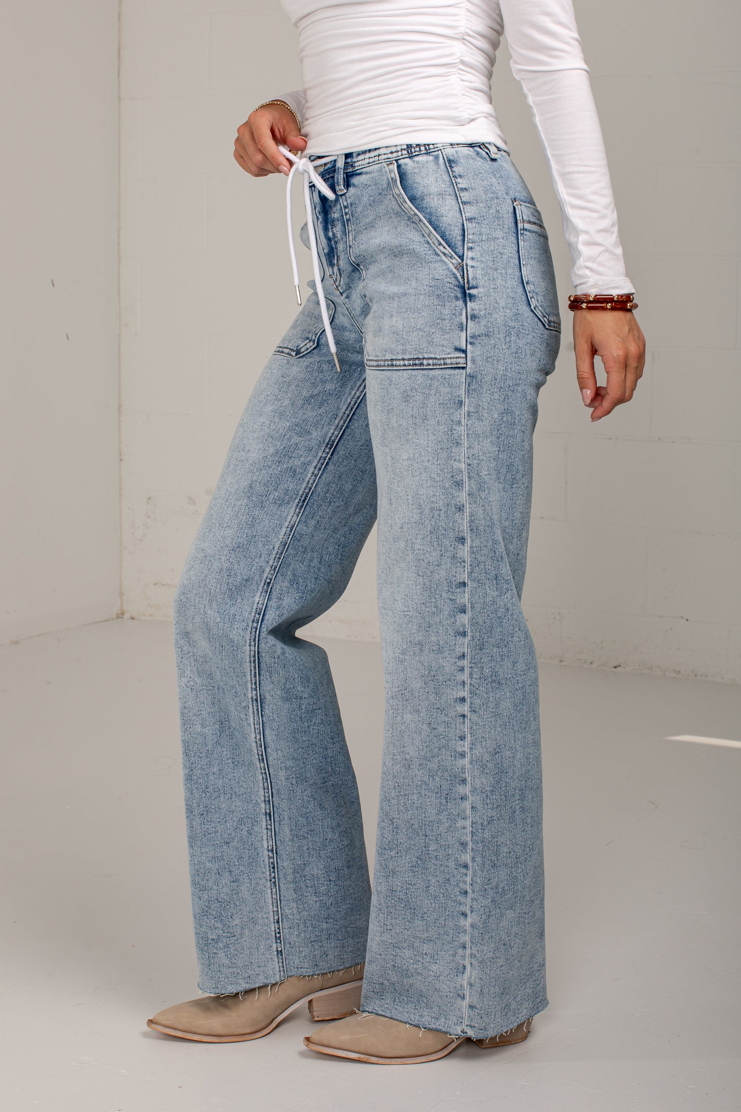 A person wearing light-wash wide-leg denim jeans with a white drawstring waist, paired with a white long-sleeve top and tan heeled shoes, standing against a plain light gray wall.
