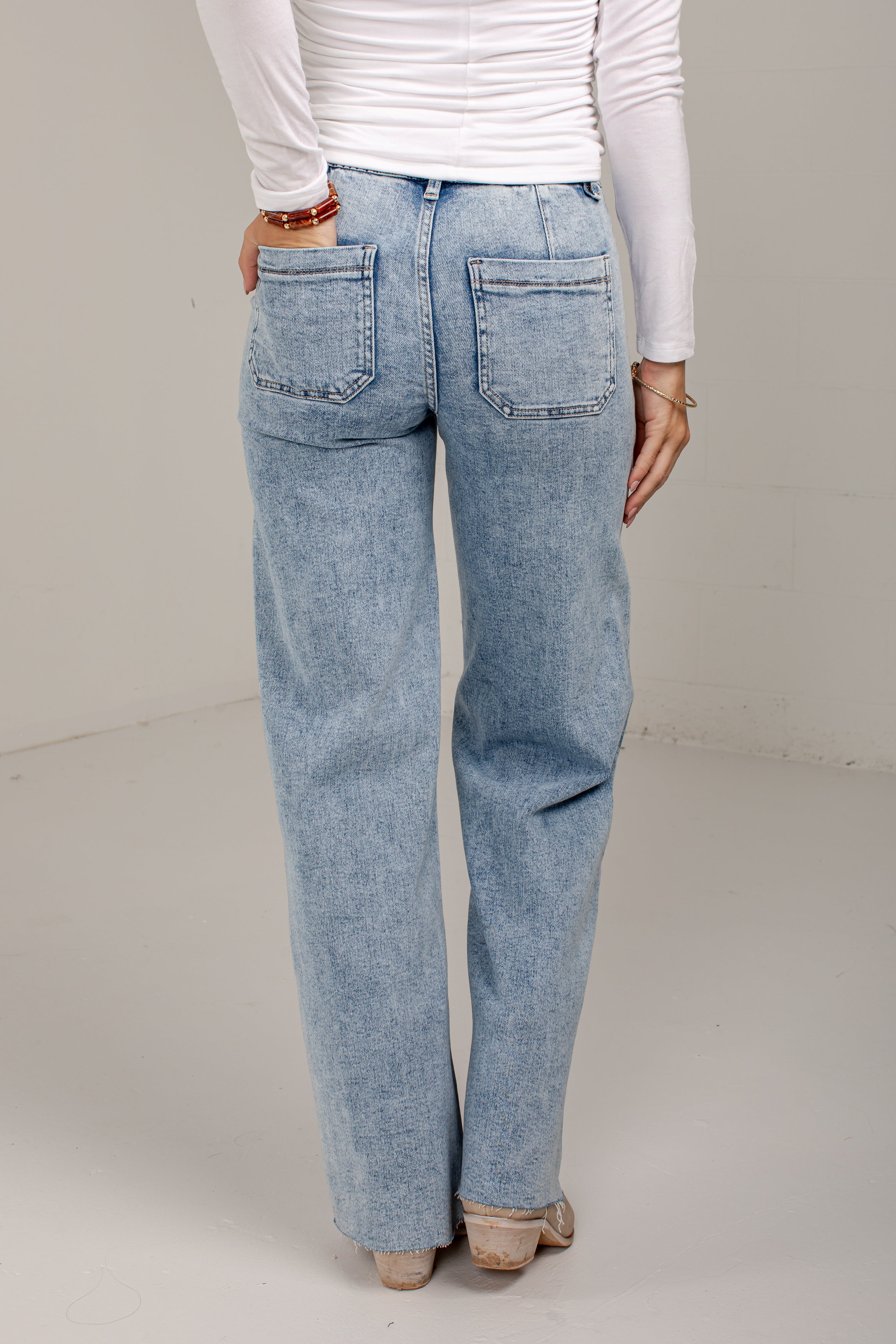 A person shown from the back wearing light-wash blue jeans, a white long-sleeve top, and beige shoes in a plain indoor studio setting.
