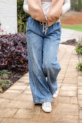 Person wearing a beige vest, white shirt, blue jeans, and white sneakers on a paved walkway.