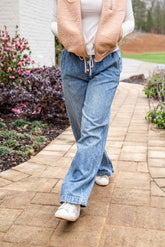 Person wearing a beige vest, white shirt, blue jeans, and white shoes on a paved walkway with plants and flowers in the background.