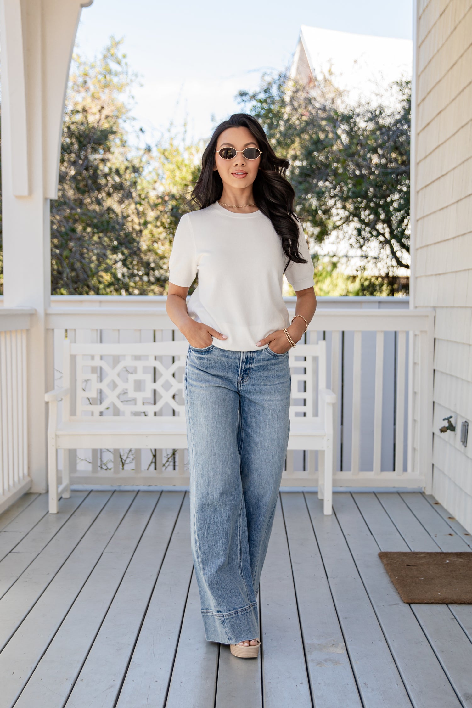 Woman standing on a porch wearing a white top and blue jeans.