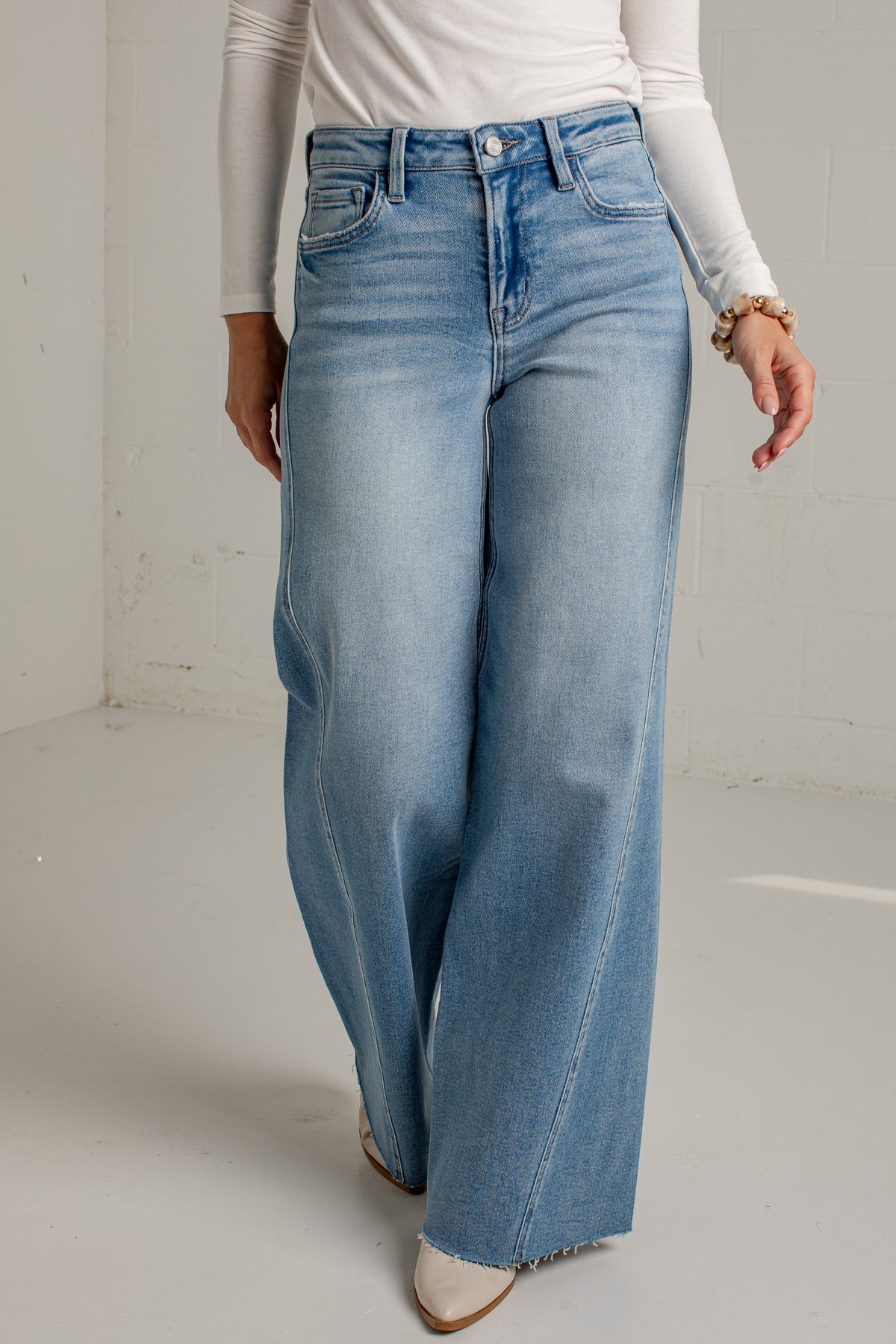 A person wearing light blue wide-leg denim jeans with a white long-sleeve top and white pointed-toe heels, standing against a plain light gray studio wall.