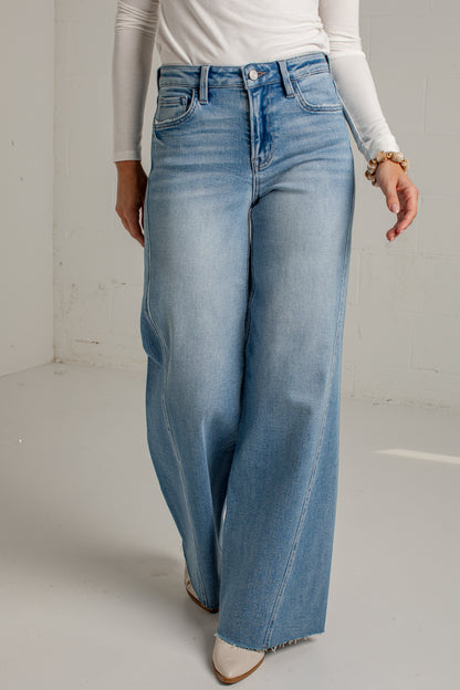 A person wearing light blue wide-leg denim jeans with a white long-sleeve top and white pointed-toe heels, standing against a plain light gray studio wall.