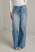 A model wearing a pair of light-wash blue wide-leg jeans, paired with a white long-sleeve top and white pointed-toe heels, standing against a white brick wall.