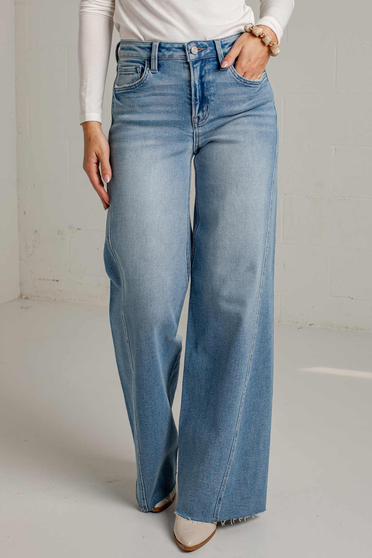 A person wearing light blue wide-leg denim jeans with a white long-sleeve top and white pointed-toe heels, standing against a light gray wall.