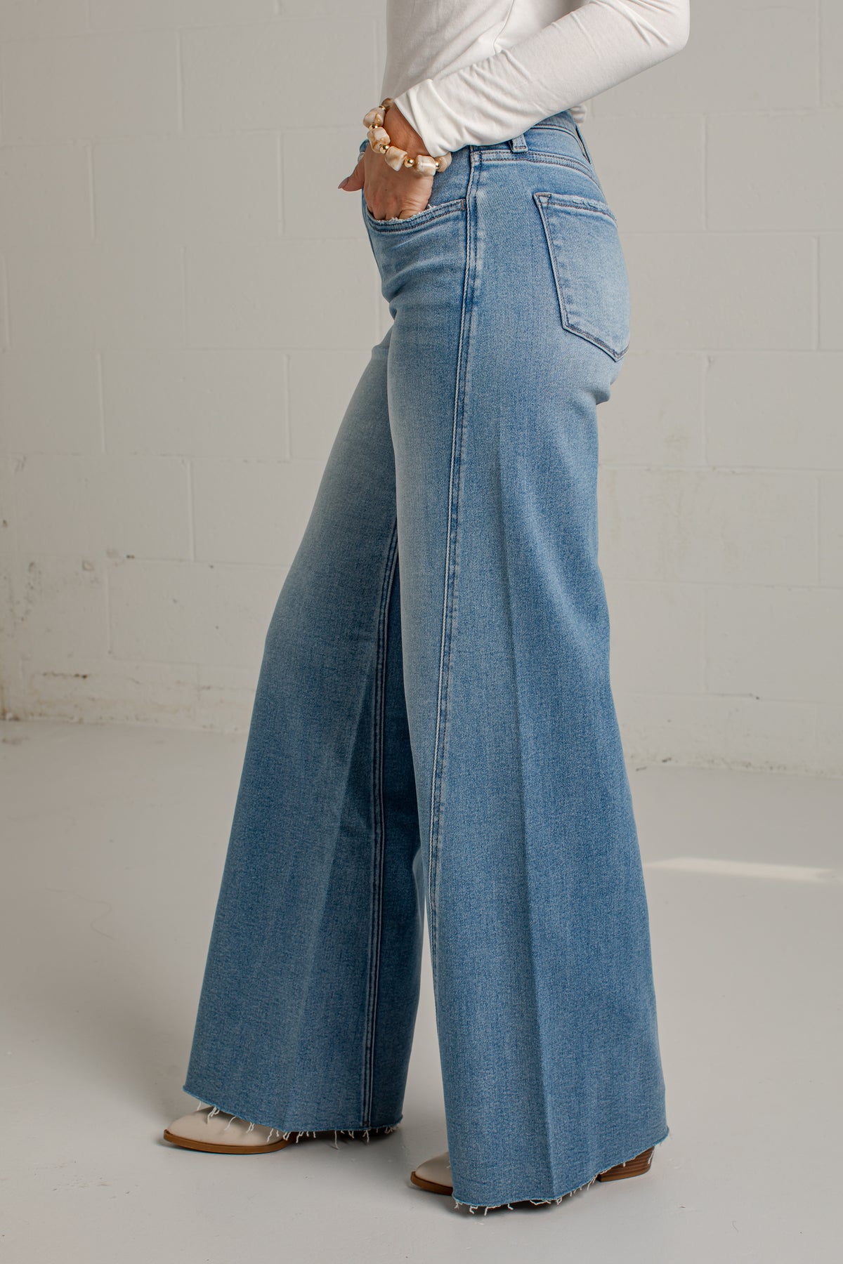 A person wearing light blue wide-leg denim jeans with a white long-sleeve top and tan sandals, standing against a light gray brick wall.