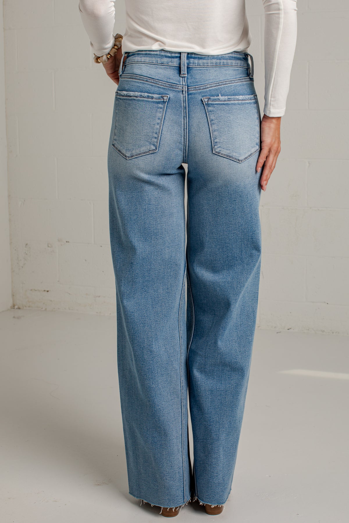 A person shown from the back wearing light blue wide-leg jeans with a white long-sleeve top, standing against a light gray wall.
