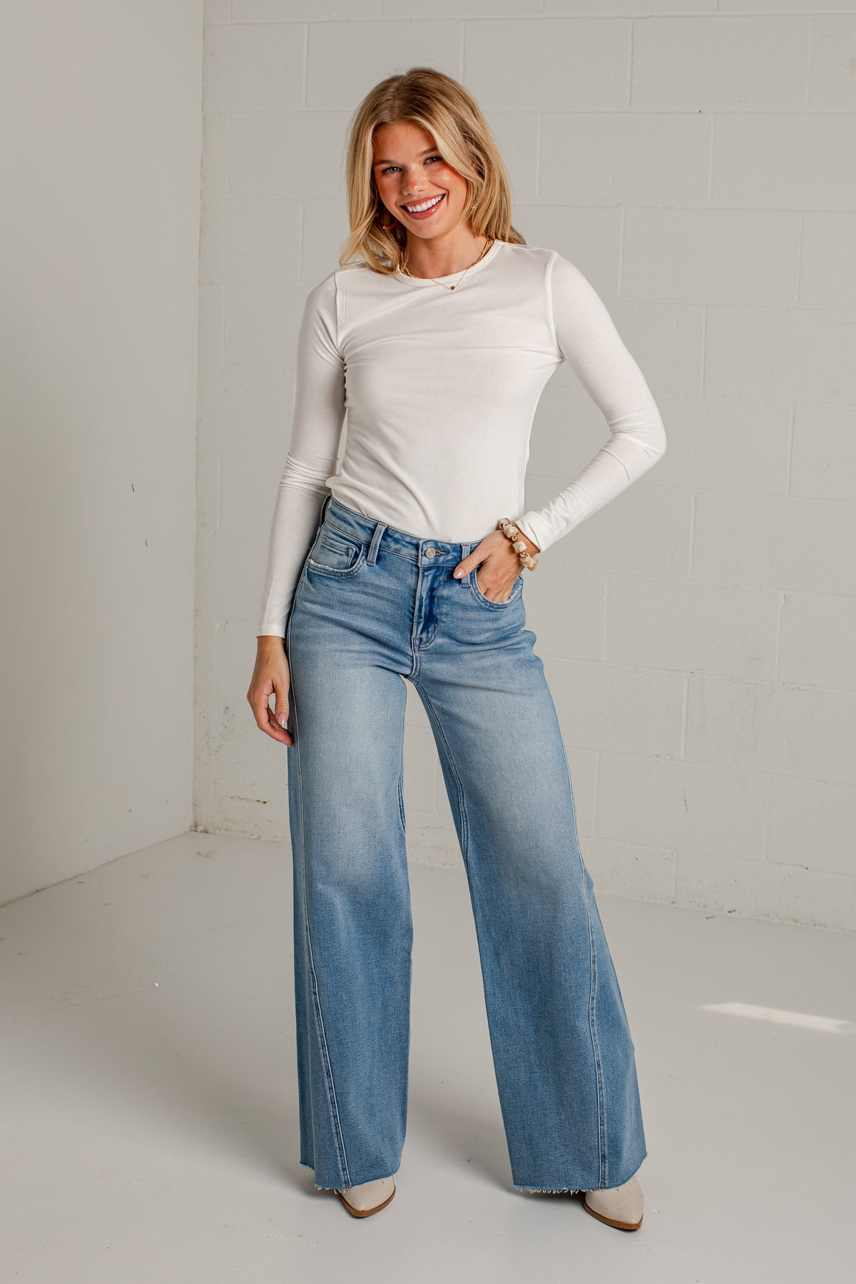 A woman modeling a white long-sleeve ribbed top and light blue wide-leg jeans in a bright, minimalist studio setting.