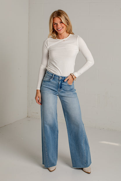 A woman modeling a white long-sleeve ribbed top and light blue wide-leg jeans in a bright, minimalist studio setting.