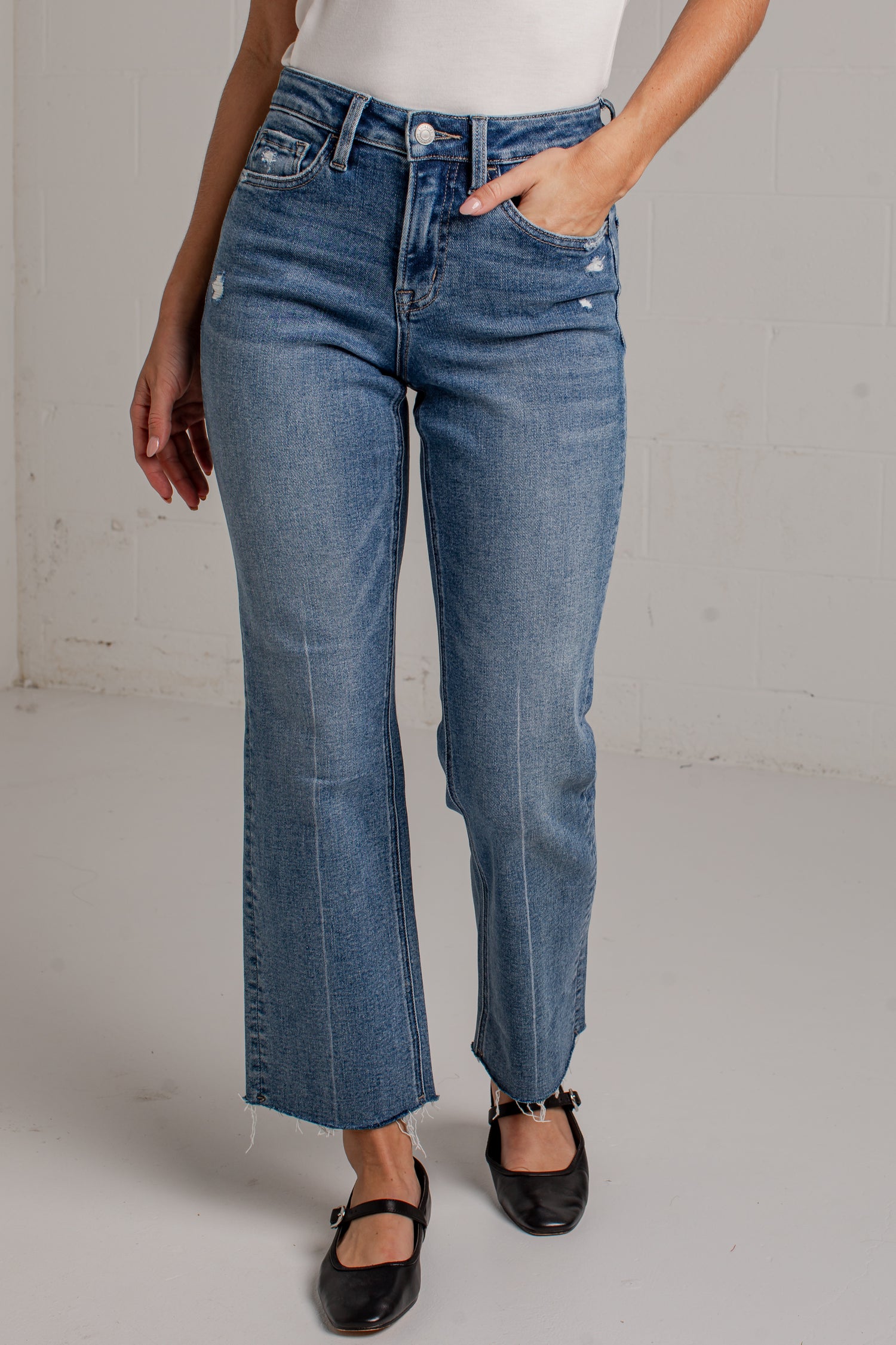 A person wearing medium-wash blue denim jeans with a straight-to-quarter leg fit, paired with a white top and black Mary Jane shoes, standing against a light gray wall.