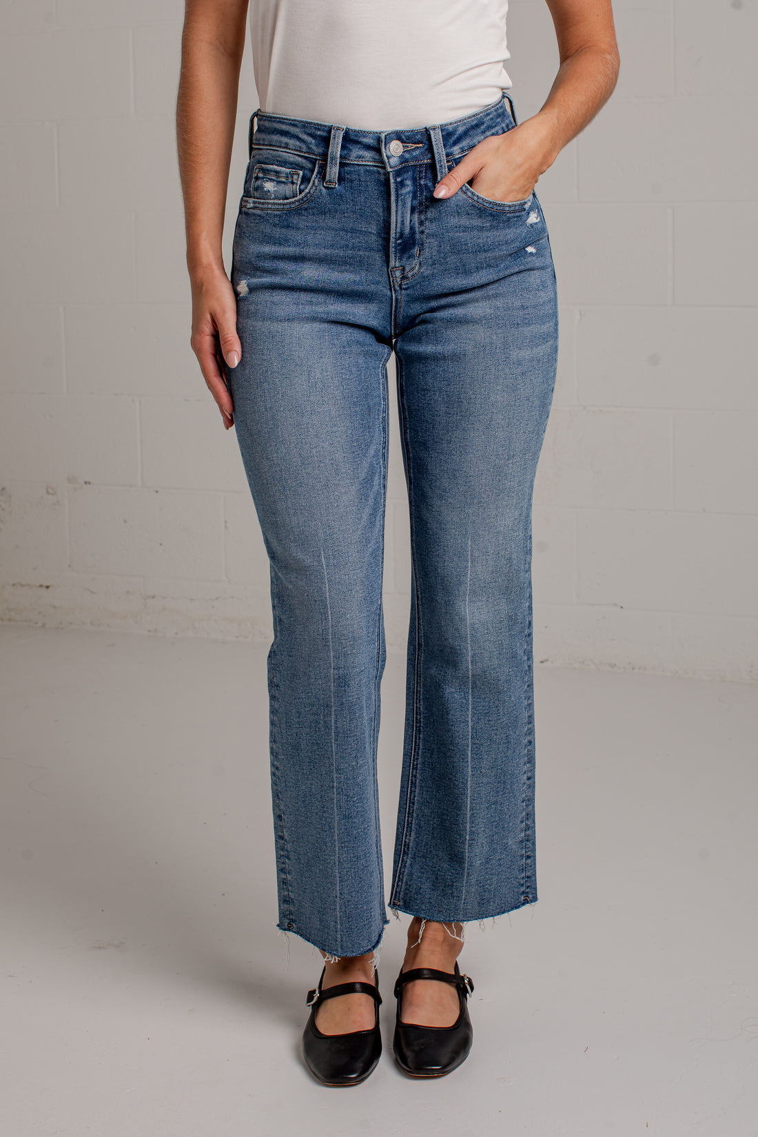 A woman modeling a white short-sleeve scoop-neck top and blue cropped wide leg jeans with black flat shoes, standing against a white brick wall.