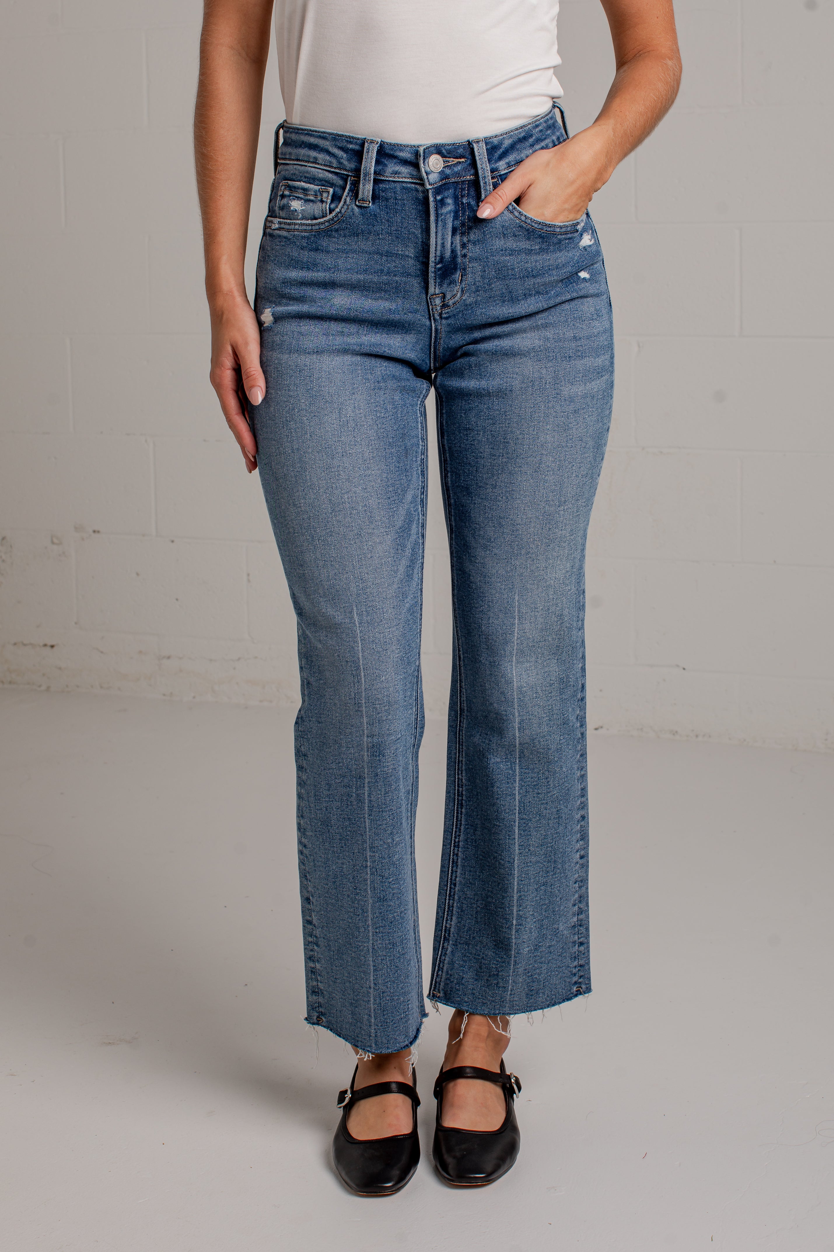 A woman modeling a white short-sleeve scoop-neck top and blue cropped wide leg jeans with black flat shoes, standing against a white brick wall.