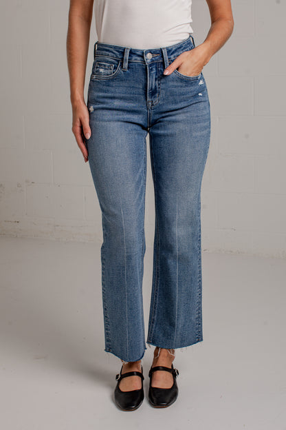 A woman modeling a white short-sleeve scoop-neck top and blue cropped wide leg jeans with black flat shoes, standing against a white brick wall.