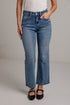 A woman modeling a white short-sleeve scoop-neck top and blue cropped wide leg jeans with black flat shoes, standing against a white brick wall.