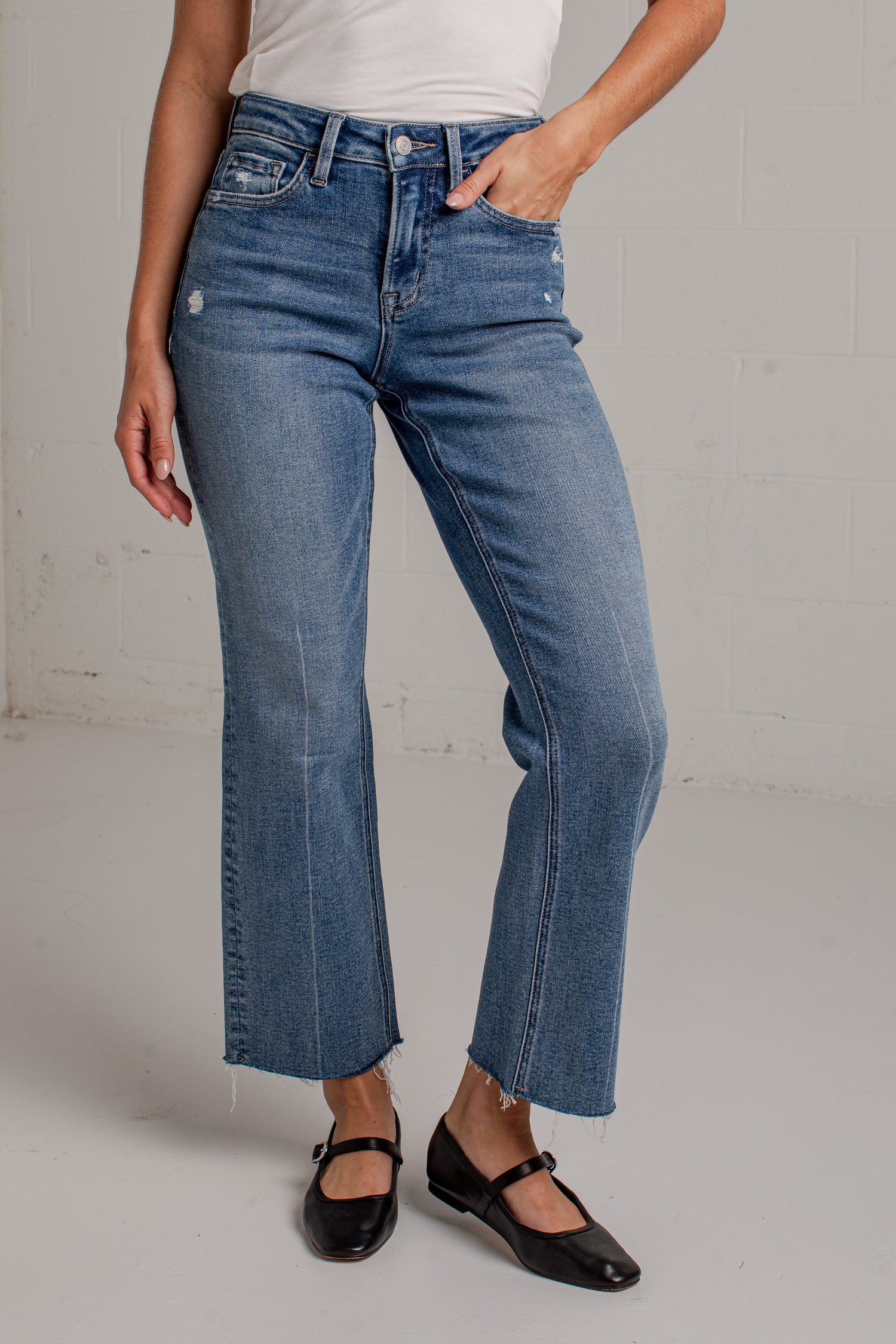 A woman modeling a white short-sleeve scoop-neck top and blue cropped wide leg jeans with black flat shoes, standing against a white brick wall.