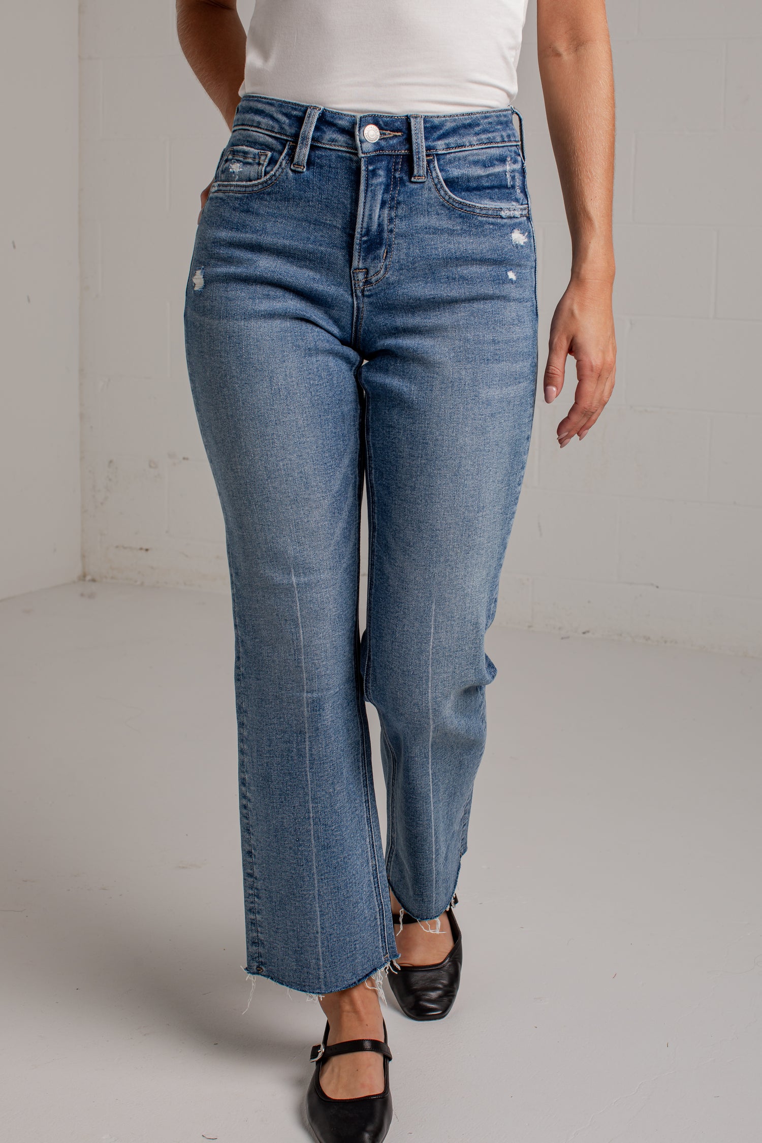 A woman modeling a white short-sleeve scoop-neck top and blue cropped wide leg jeans with black flat shoes, standing against a white brick wall.