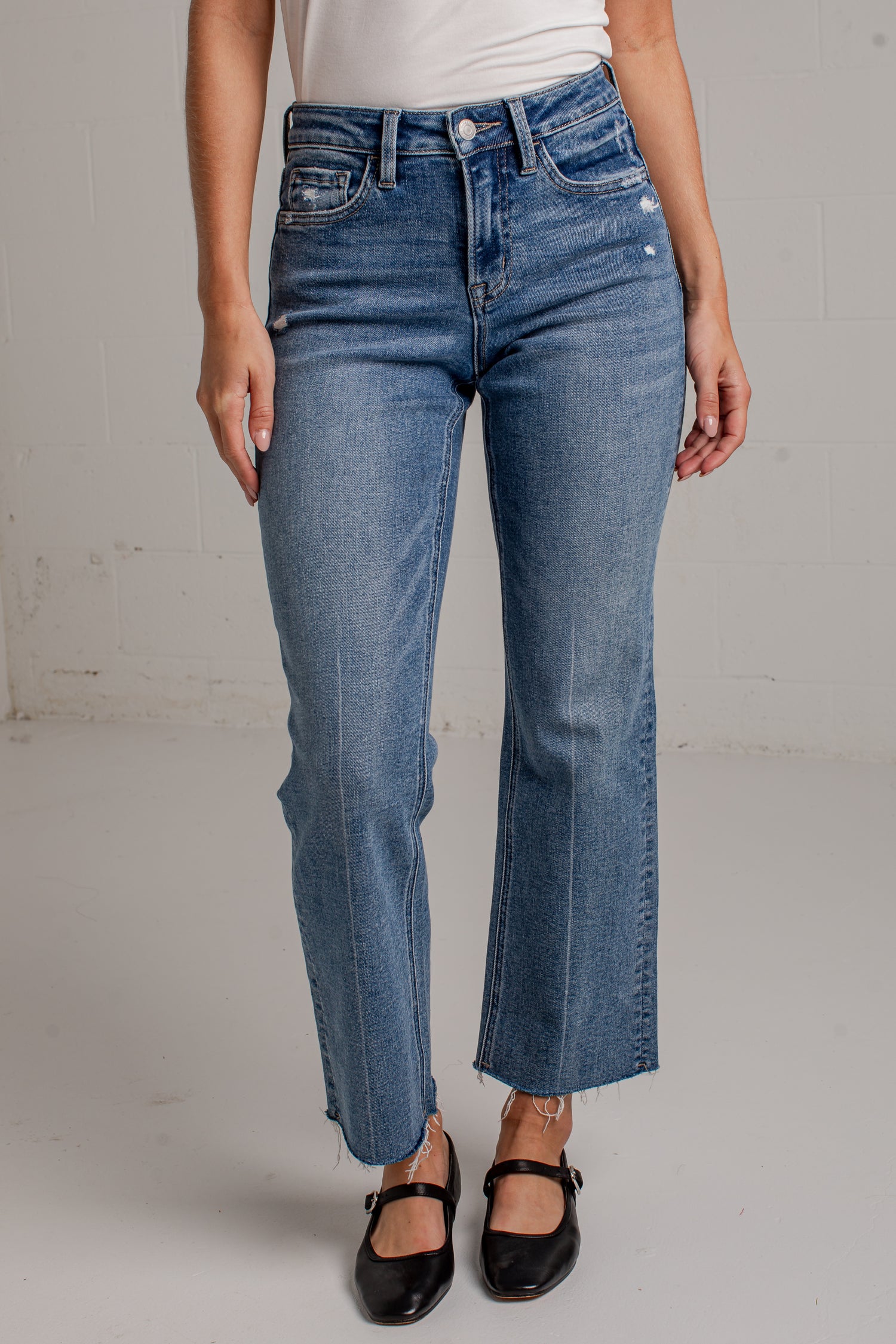 A woman modeling a white short-sleeve scoop-neck top and blue cropped wide leg jeans with black flat shoes, standing against a white brick wall.