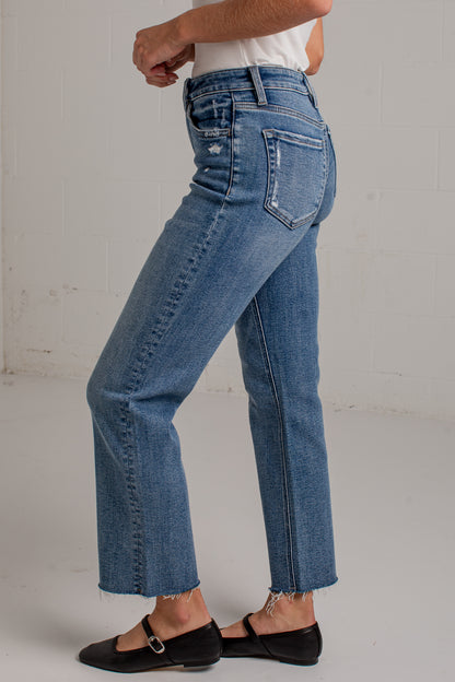A person wearing medium-wash blue denim jeans with a straight-to-crew leg, frayed hems, and distressed details on the back pockets, paired with black flat shoes against a white brick wall background.