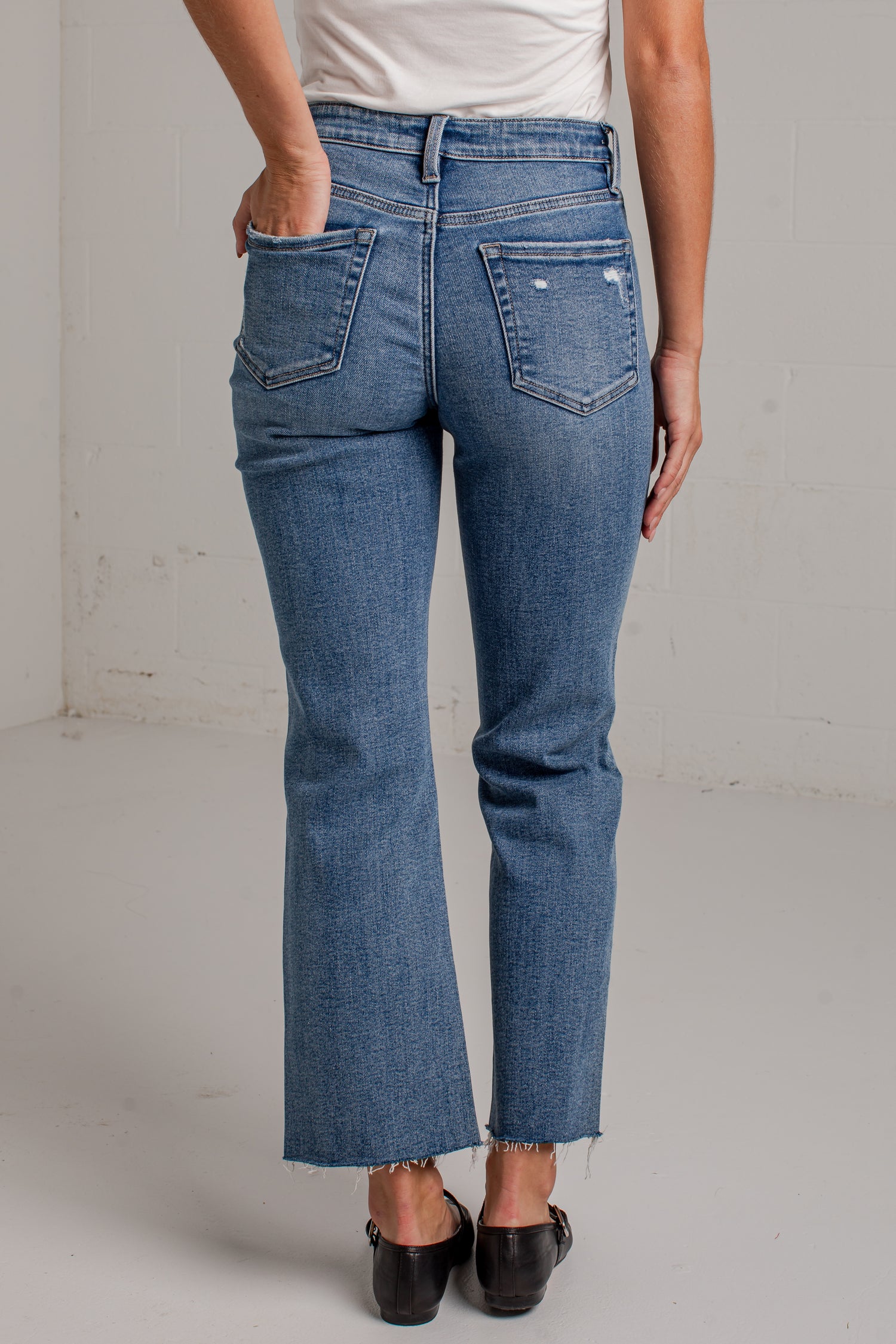 A woman modeling a white short-sleeve scoop-neck top and blue cropped wide leg jeans with black flat shoes, standing against a white brick wall.