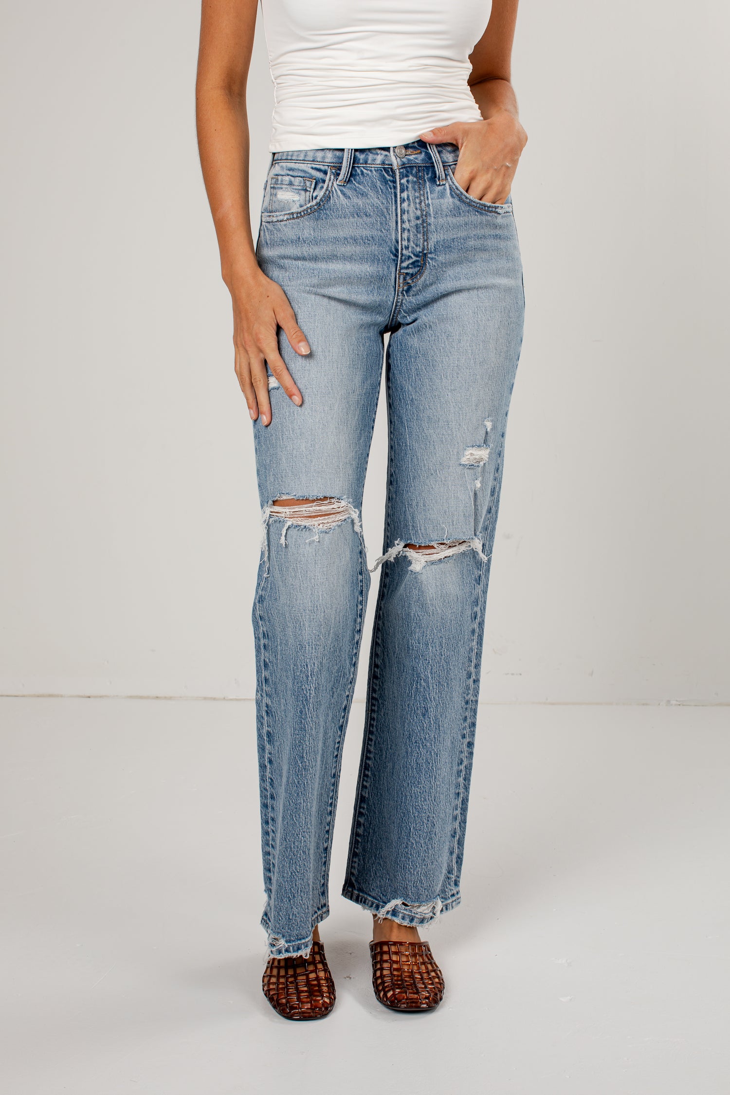 A model wearing light-wash blue distressed jeans with ripped details on the thighs and knees, paired with a white top and brown woven flat shoes, standing against a light gray studio background.