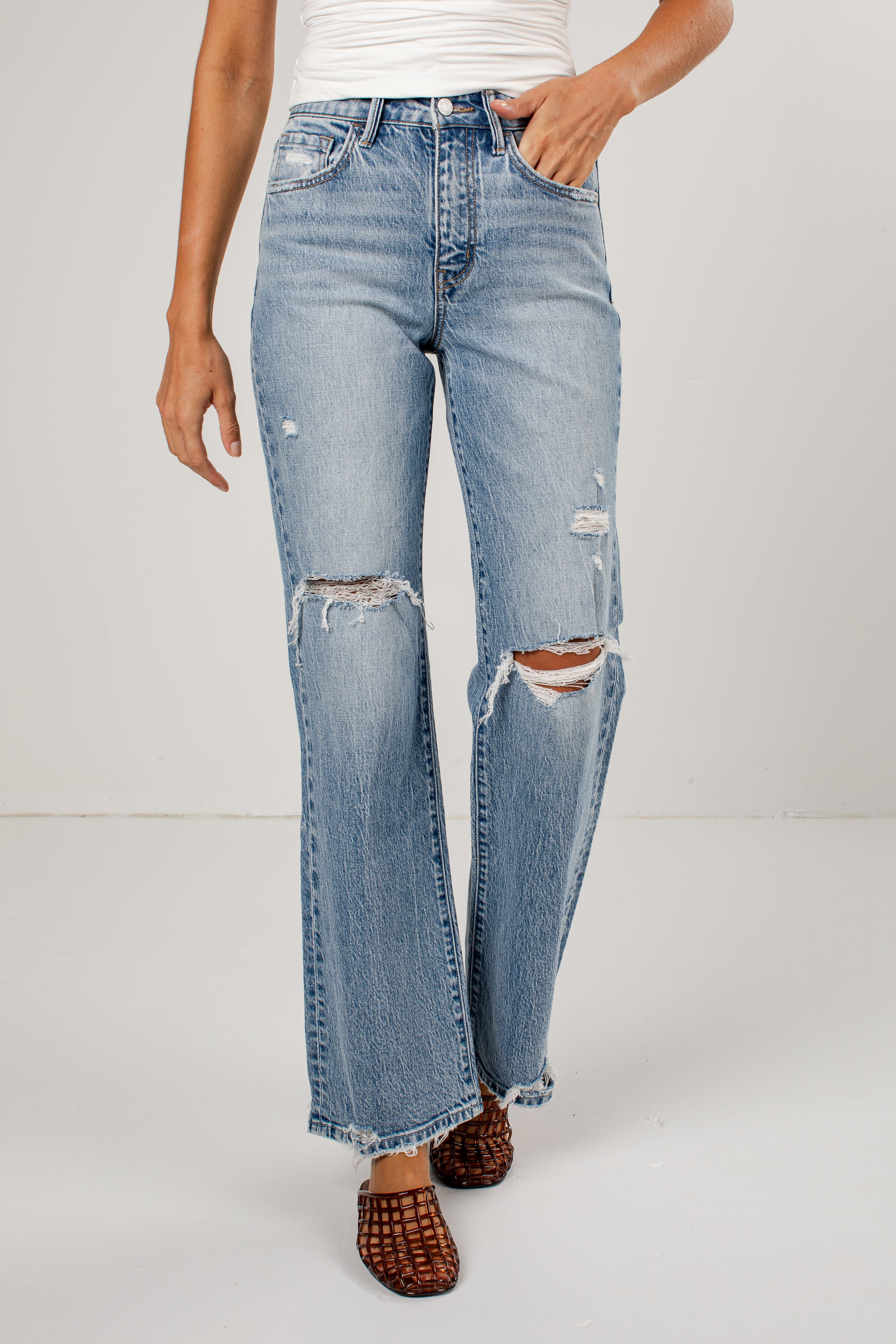 A person wearing medium-wash blue distressed jeans with ripped knees and a straight-leg fit, paired with brown woven sandals against a plain light gray studio background.