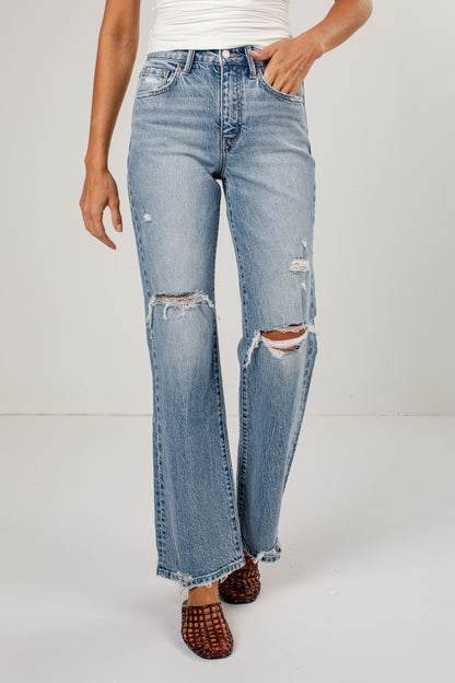 A person wearing medium-wash blue distressed jeans with ripped knees and a straight-leg fit, paired with brown woven sandals against a plain light gray studio background.