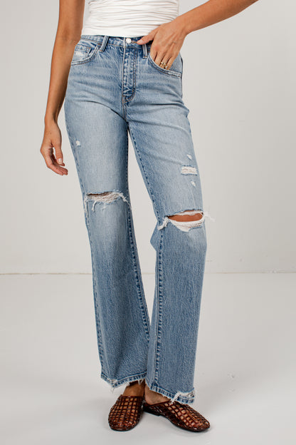 A person wearing light-wash blue distressed jeans with ripped knees and a straight-leg fit, paired with brown woven flat shoes against a plain light gray studio background.