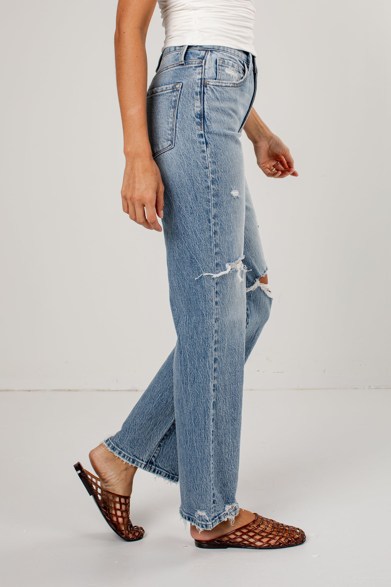 A person wearing light-wash blue distressed jeans with a straight-to-wide leg, paired with brown woven mule sandals against a plain light gray studio background.