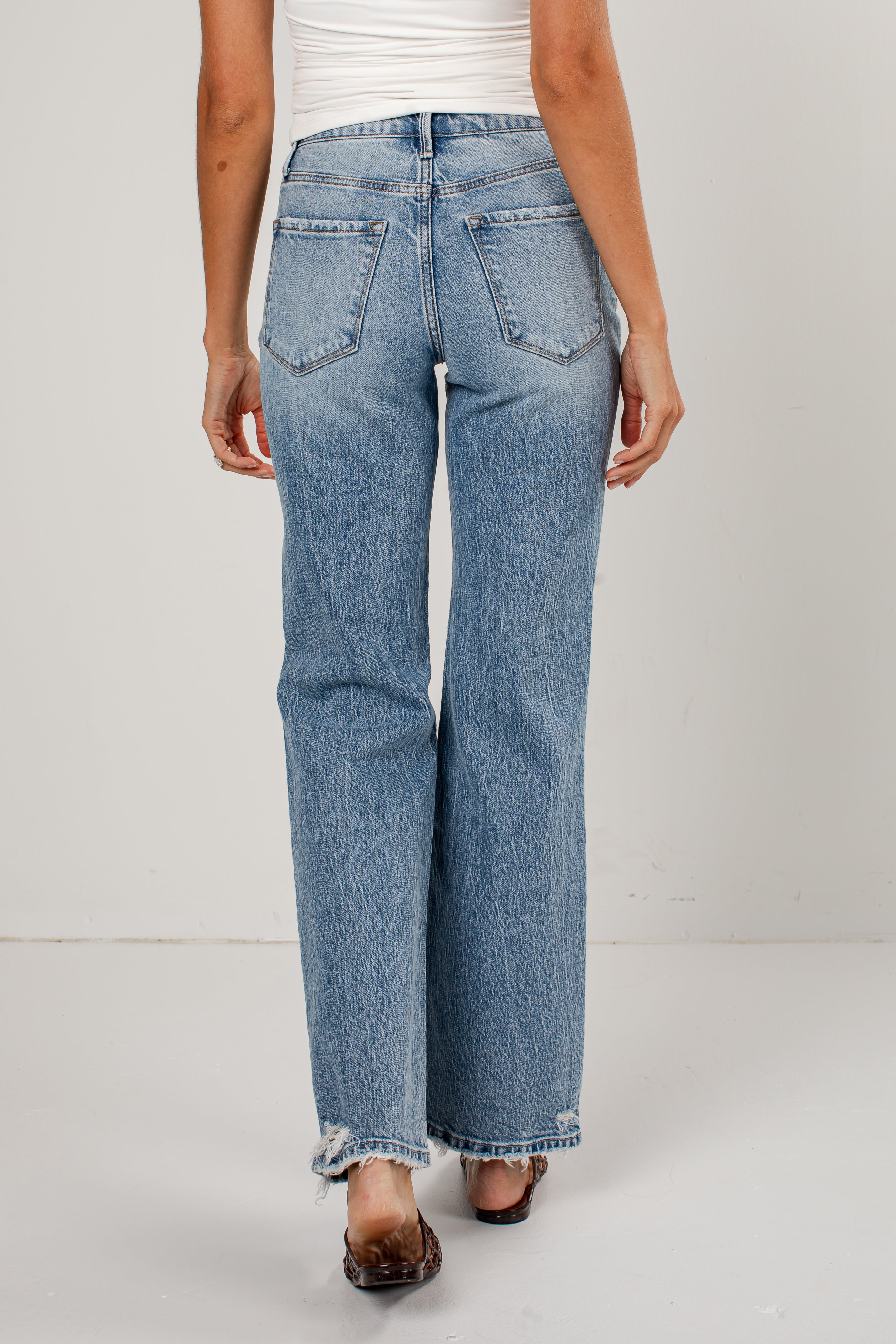 A person shown from the back wearing light-wash blue jeans with a straight-to-wide leg fit, cuffed hems, and raw frayed detailing. The model is also wearing a white top and brown flat sandals, standing against a plain light gray studio background.