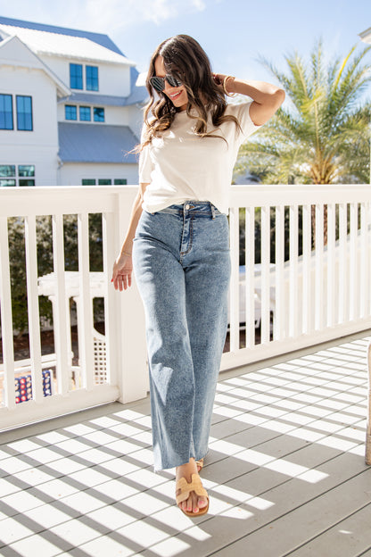 Woman standing on a deck wearing a white top and blue jeans, with a house and palm tree in the background.