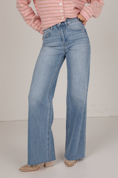A person wearing light blue high-waisted wide-leg jeans, a pink and white striped long-sleeve top, and beige pointed-toe heels, standing against a plain light beige studio background.