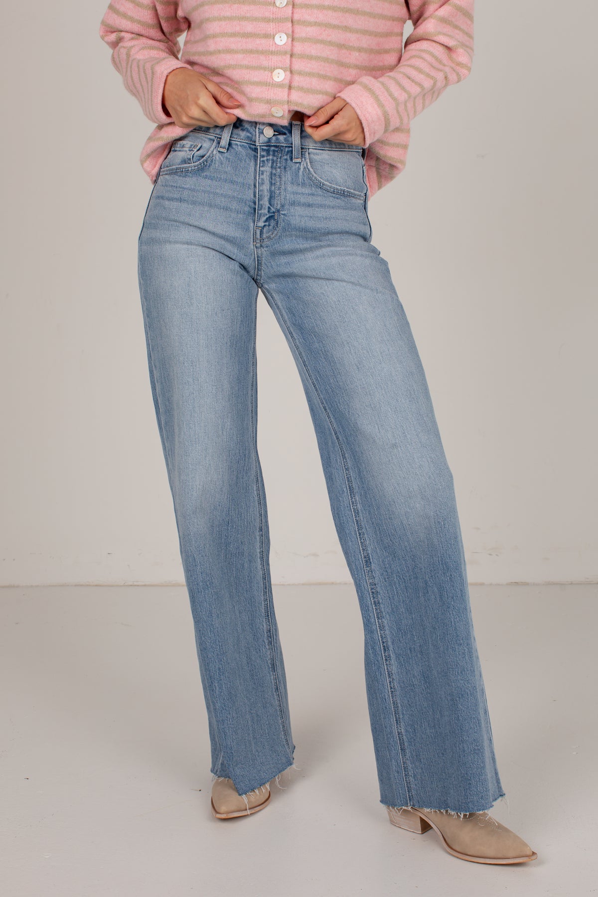 A person wearing light-wash blue wide-leg jeans, a pink and white striped long-sleeve top, and beige pointed-toe flats, standing against a plain light gray studio background.
